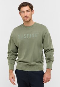 Sweatshirt vert olive en mélange de coton doux, avec des poignets et un ourlet côtelés, arborant le logo "MUSTANG" en relief légèrement sur le devant.