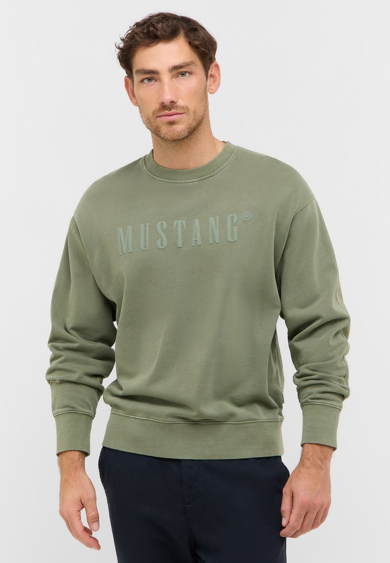 Sweatshirt vert olive en mélange de coton doux, avec des poignets et un ourlet côtelés, arborant le logo "MUSTANG" en relief légèrement sur le devant.