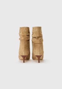 Stivaletti alla caviglia in suede beige con un design arricciato e un tacco in legno a punta. Texture liscia senza decorazioni visibili.
