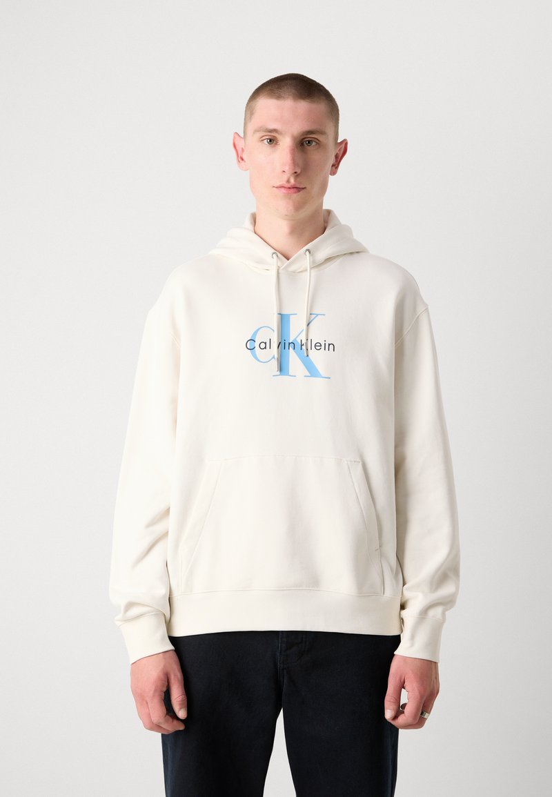 Felpa con cappuccio di colore crema in tessuto morbido, con tasca frontale e un logo blu con la scritta "Calvin Klein".