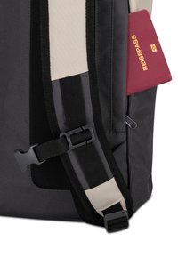 Zaino nero e grigio con un pannello beige, cintura regolabile con fibbia in plastica e un passaporto marrone scuro parzialmente visibile.