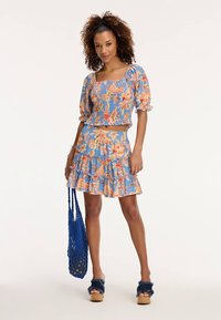 Top albastru cu imprimeu floral și mâneci bufante, asortat cu o fustă plisată, cu modele portocalii și galbene. Modelul ține o geantă albastră din plasă.