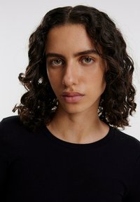 Porträt einer Person mit lockigem, dunklem Haar, die ein schwarzes, geripptes Kurzarmoberteil trägt. Der Hintergrund ist neutral und lenkt den Fokus auf die Gesichtszüge.