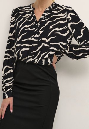 Frau trägt eine schwarz-weiß zebragestreifte Bluse, die in einen enganliegenden schwarzen Rock gesteckt ist, Hand in der Hüfte, neutraler Hintergrund.