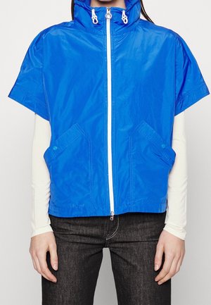 Osoba nosząca jasnoniebieską kurtkę na krótki rękaw z zamkiem błyskawicznym i kieszeniami, na białej koszuli z długimi rękawami i ciemnych dżinsach.