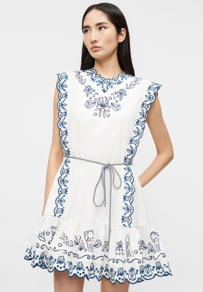 Νεαρή γυναίκα που φοράει λευκό αμάνικο φόρεμα με μπλε κεντήματα λουλουδιών, κυματιστά άκρα και λεπτή ζώνη δεμένη στη μέση.