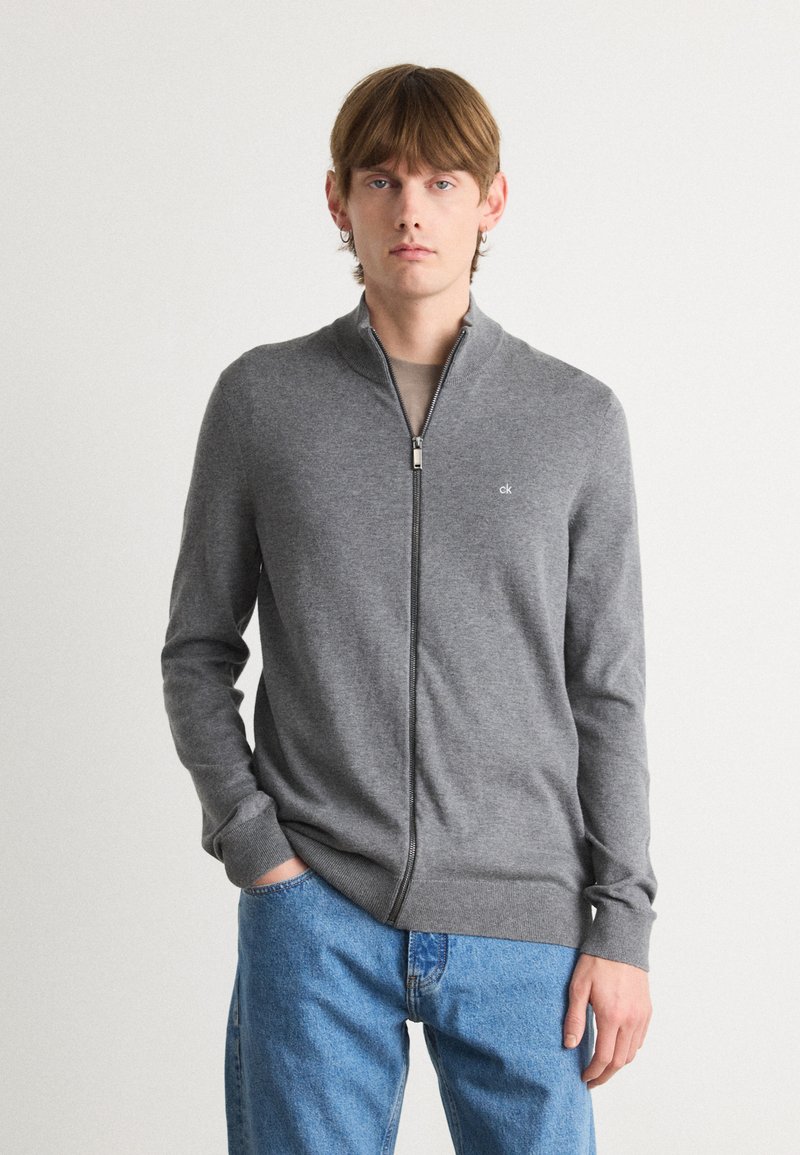 Sweat-shirt zippé gris en tissu doux, avec un col montant et des poignets côtelés. Petit logo sur la poitrine gauche. Porté avec un jean bleu.