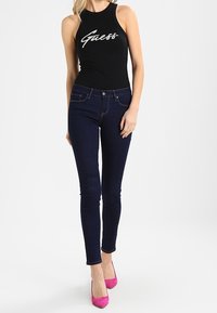 Haut noir sans manches avec le logo "Guess", associé à un jean skinny bleu foncé et des chaussures à talons hauts roses. Fond neutre.