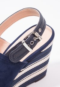 Sandale compensée bleu marine avec bride de cheville en cuir ajustable et boucle argentée, dotée d'un talon plateforme tissé rayé bleu marine et blanc.