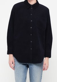 Personne portant une chemise en velours côtelé noir à boutons avec manches longues et poche poitrine, associée à un jean bleu clair.