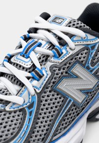 Pilkas tinklinis bėgimo batelis su mėlynais ir sidabriniais akcentais, turintis raštuotą dizainą ir baltas raišteles. Šone matomas ryškus „N“ logotipas.