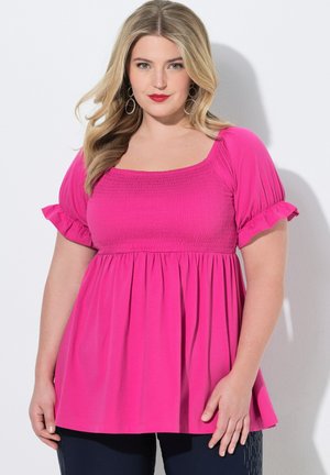 Mujer con cabello largo y rubio, lleva pendientes de aro plateados y una blusa fruncida de color rosa brillante con mangas cortas con volantes, de pie frente a un fondo blanco.