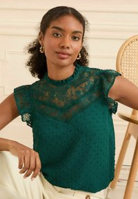 Tealfärgad blus med spetsdetaljer, texturerat prickmönster, volangkrage och korta ärmar. Modellen är i en neutral miljö med en stol.