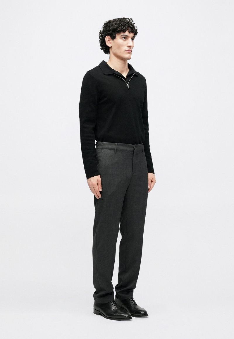 Black zip-neck sweater, gray tailored trousers, and black dress shoes. Curly hair, standing pose, neutral background.