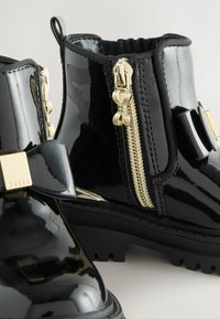 Primer plano de botas de tobillo negras brillantes con detalles de cremallera dorada y un lazo decorativo en el costado, sobre un fondo claro.