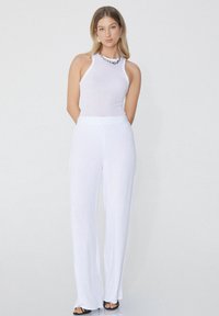 Bílý žebrovaný halter top s kulatým výstřihem v kombinaci s bílými širokými kalhotami s vysokým pasem. Obojí je vyrobeno z lehkého materiálu.