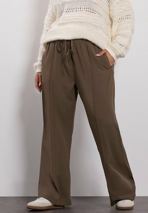 Person wearing loose brown drawstring pants with pockets, a cream knit sweater, and white sneakers against a plain wall.