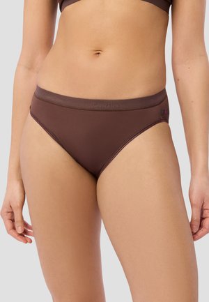 Frau trägt braune High-Cut-Slips und passenden BH, gezeigt vom mittleren Oberkörper bis zu den oberen Oberschenkeln vor eintönigem Hintergrund.