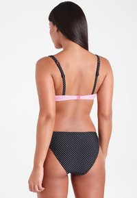 Schwarzer Bikini mit weißen Pünktchen und einem pinken Gingham-Träger, der ein schlichtes Design und einen verstellbaren Verschluss auf der Rückseite aufweist. Das Material wirkt glatt und dehnbar.