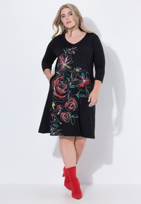 Robe noire avec des manches 3/4, ornée de broderies florales multicolores sur le devant et de poches latérales. Associée à des bottes à talons rouges.