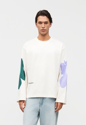 Jonge man staand, naar voren kijkend, draagt een wit shirt met lange mouwen met grote groene en paarse bloemenprints op de mouwen en lichte blauwe spijkerbroek.