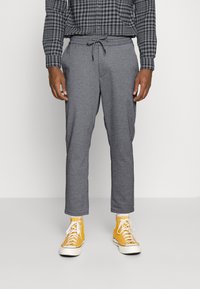 Hombre vestido con pantalones grises con cordón, zapatillas altas amarillas y una camisa de manga larga a cuadros en negro y gris, de pie sobre un fondo liso.