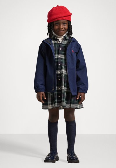 Mornarska jakna, zelena karirana obleka, rdeča pletena kapa, nogavice do kolen mornarske barve in sijoči črni čevlji. Obleka združuje različne teksture in barve.