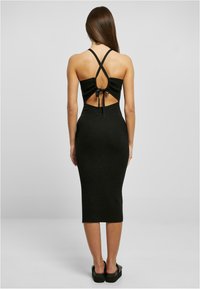 Abito midi nero a costine con spalline incrociate, dettaglio della schiena scoperta con fiocco e silhouette aderente. Scarpe nere piatte e spesse.