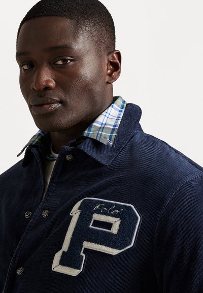 Navy corduroy jacket with a plaid collared shirt underneath, featuring a white embroidered 'P' logo on the front.