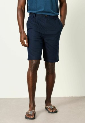 Mees, kes kannab tumesiniseid lühikesi pükse, sinist t-särki ja mitmevärvilisi sandaale, seisab ühe käega taskus tavalise tausta ees.