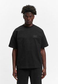 Schwarzes Baumwoll-T-Shirt mit kurzen Ärmeln, legerer Schnitt, gesticktes Logo auf der Brust und einer glatten Textur. Kombiniert mit schwarzen Hosen.