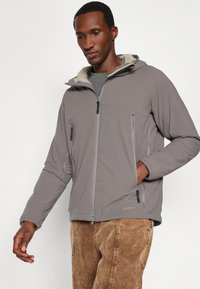 Veste zippée grise avec une capuche en fausse fourrure, deux poches zippées et des poignets élastiques. Portée avec un pantalon en velours côtelé marron. Texture de tissu lisse.