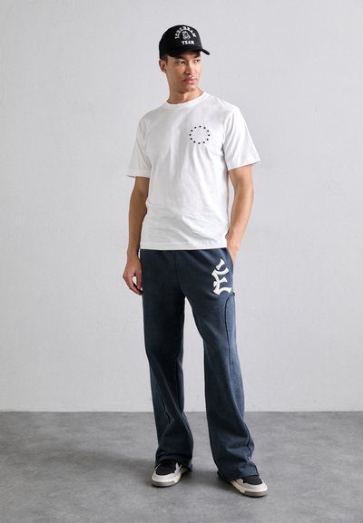 Vit T-shirt med kort ärm och svart stjärntryck, matchad med marinblåa sweatpants med vit logotyp. En svart keps fulländar looken.