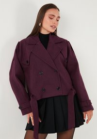 Dvojno zapenjalna, skrajšana jakna v temno vijoličnem volnenem mešanici z belimi gumbi, širokimi reverji in pasovno vezavo. Poudarjena z oblikovanimi rokavi.