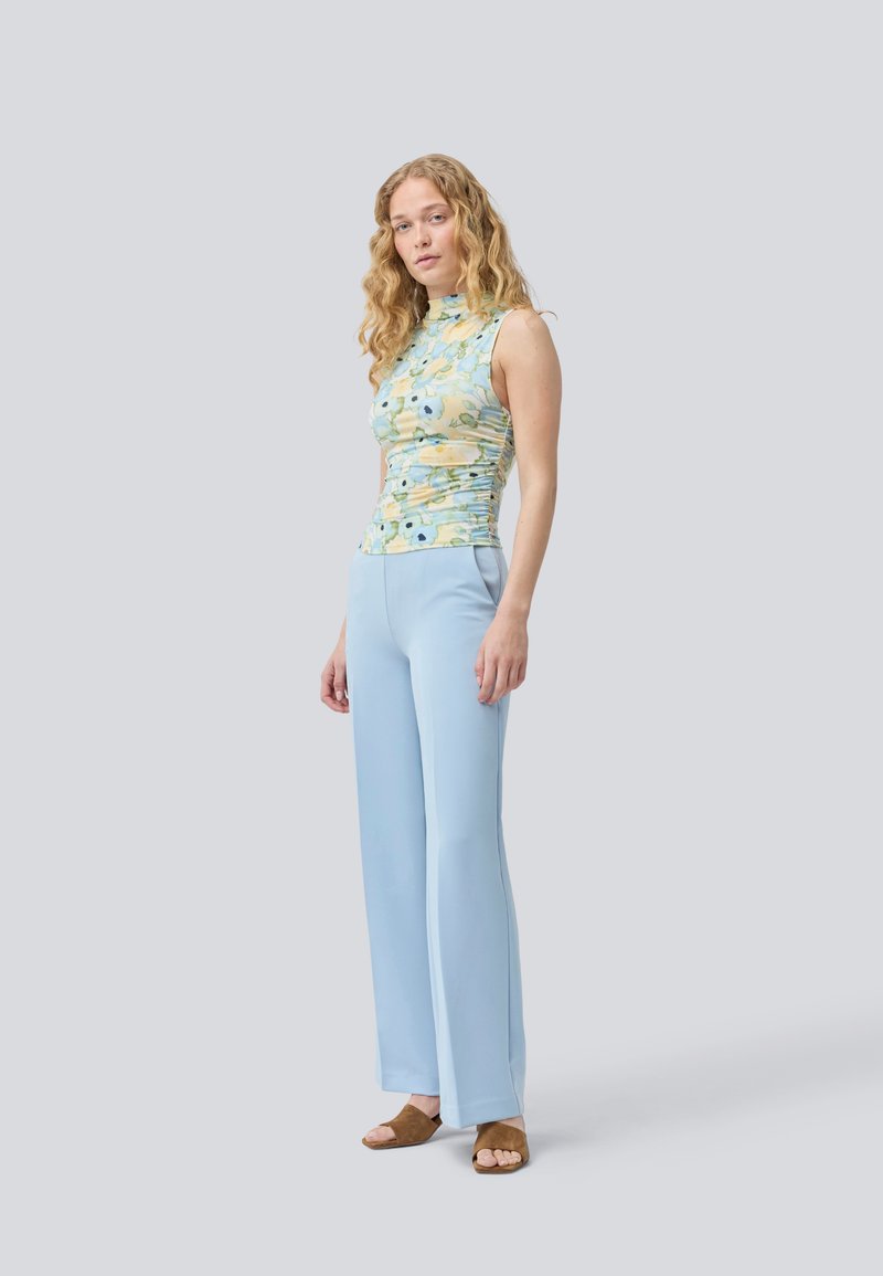 Floral-patterned, sleeveless top with a high neck, paired with light blue, flared trousers. Brown suede slide sandals complete the look.