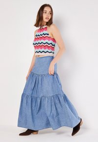 Top halter de ganchillo con patrón de zigzag multicolor. Falda larga de mezclilla con volantes en capas, tono azul claro, combinada con botas de tacón marrones.