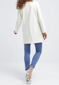 Lång, vit kappa i ett mjukt material, samspelet med blå denimjeans och silverfärgade högklackade skor. Vy från baksidan.