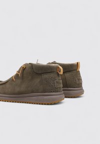 Zapatos de ante oliva con suela de goma texturizada, que presentan un cuello de pana y acentos en marrón claro contrastantes. Diseño flexible para mayor comodidad.