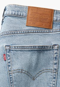 Ljusblå denimjeans med en brun läderpatch vid midjan. Har signaturstickning, en röd etikett på vänstra fickan och nitar.