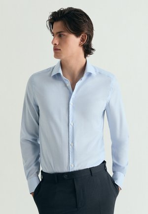 Joven con pelo oscuro que lleva una camisa azul claro abotonada y pantalones negros, mirando hacia la izquierda con las manos en los bolsillos.