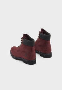 Burgunderfarbene Wildleder-Stiefeletten mit einem gepolsterten schwarzen Kragen, Metallösen und einer strukturierten Gummisohle, die ein Logo an der Seite aufweisen.