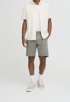 Homme portant une chemise beige à manches courtes par-dessus un t-shirt blanc, un short gris retroussé, des chaussettes blanches mi-mollet et des baskets beiges.