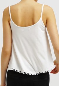 Camisola bianca con spalline sottili, schiena arrotondata e bordo con pom pom lungo l'orlo. Realizzata in tessuto morbido e leggero con una vestibilità ampia.