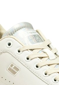 Sneaker bianco in pelle con linguetta imbottita argentata lucida, lacci crema e un piccolo logo vicino all'area della punta. Presenta occhielli rotondi.