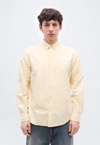 Jeune homme aux cheveux courts et bouclés, portant une chemise jaune pâle boutonnée et un jean bleu, debout devant un fond blanc.