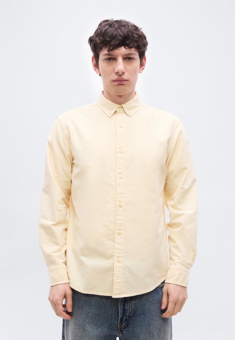 Jeune homme aux cheveux courts et bouclés, portant une chemise jaune pâle boutonnée et un jean bleu, debout devant un fond blanc.