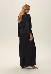 Long black satin garment with loose sleeves and a belted waist, featuring side slits and wide pants. Silver strappy shoes visible.