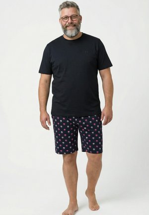 Mężczyzna z siwymi włosami i brodą, nosi okulary, czarny t-shirt i granatowe szorty z różowo-białym wzorem palm, stoi boso.