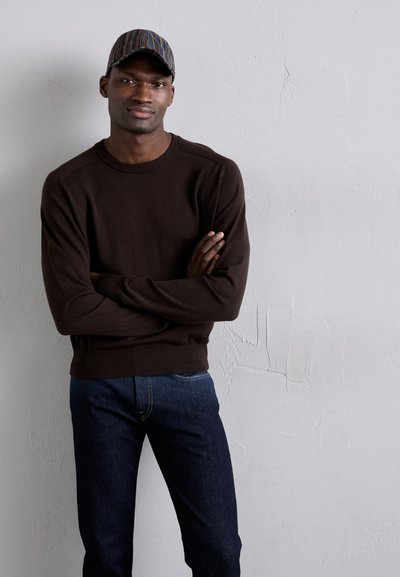 Suéter de punto marrón con puños acanalados, combinado con jeans de mezclilla azul oscuro. El modelo lleva una gorra de rayas multicolores.