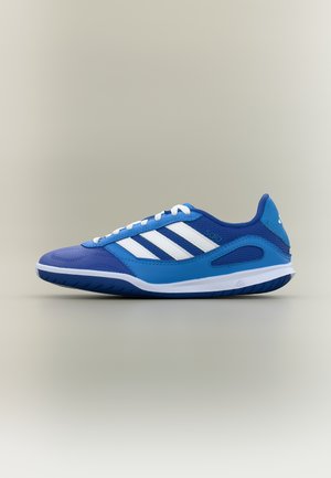 Zapatilla de fútbol sala Adidas azul y blanca con cordones blancos, tres franjas blancas en el lateral y suela texturizada sobre un fondo liso.